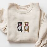 Sand crewneck sweatshirt laid flat, embroidered with a black and white tuxedo pit bull and a fawn pit bull sitting side by side with their tongues out, centered on the chest