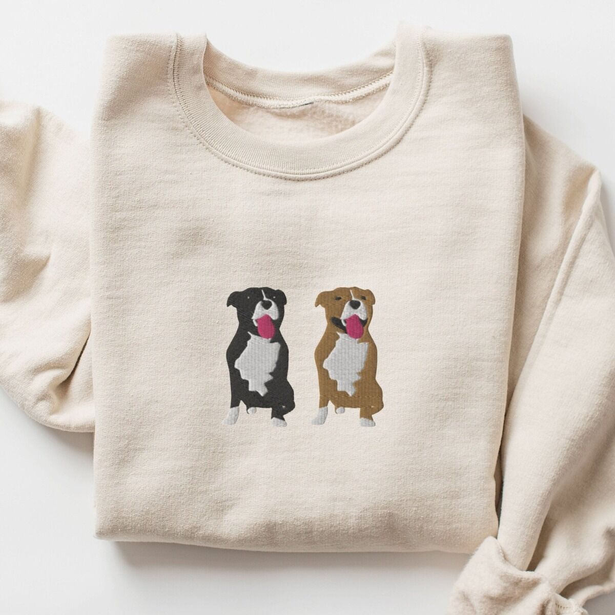 Two Pitbulls Sweatshirt – Sand Sand crewneck sweatshirt laid flat, embroidered with a black and white tuxedo pit bull and a fawn pit bull sitting side by side with their tongues out, centered on the chest
