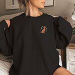 Woman sitting on a bed in a Black crewneck sweatshirt embroidered with a small golden rust vizsla sitting among white snowflakes on the left chest