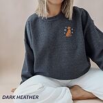 Woman sitting in white pants and a Dark Heather crewneck sweatshirt embroidered with a small golden rust vizsla sitting among white snowflakes on the left chest