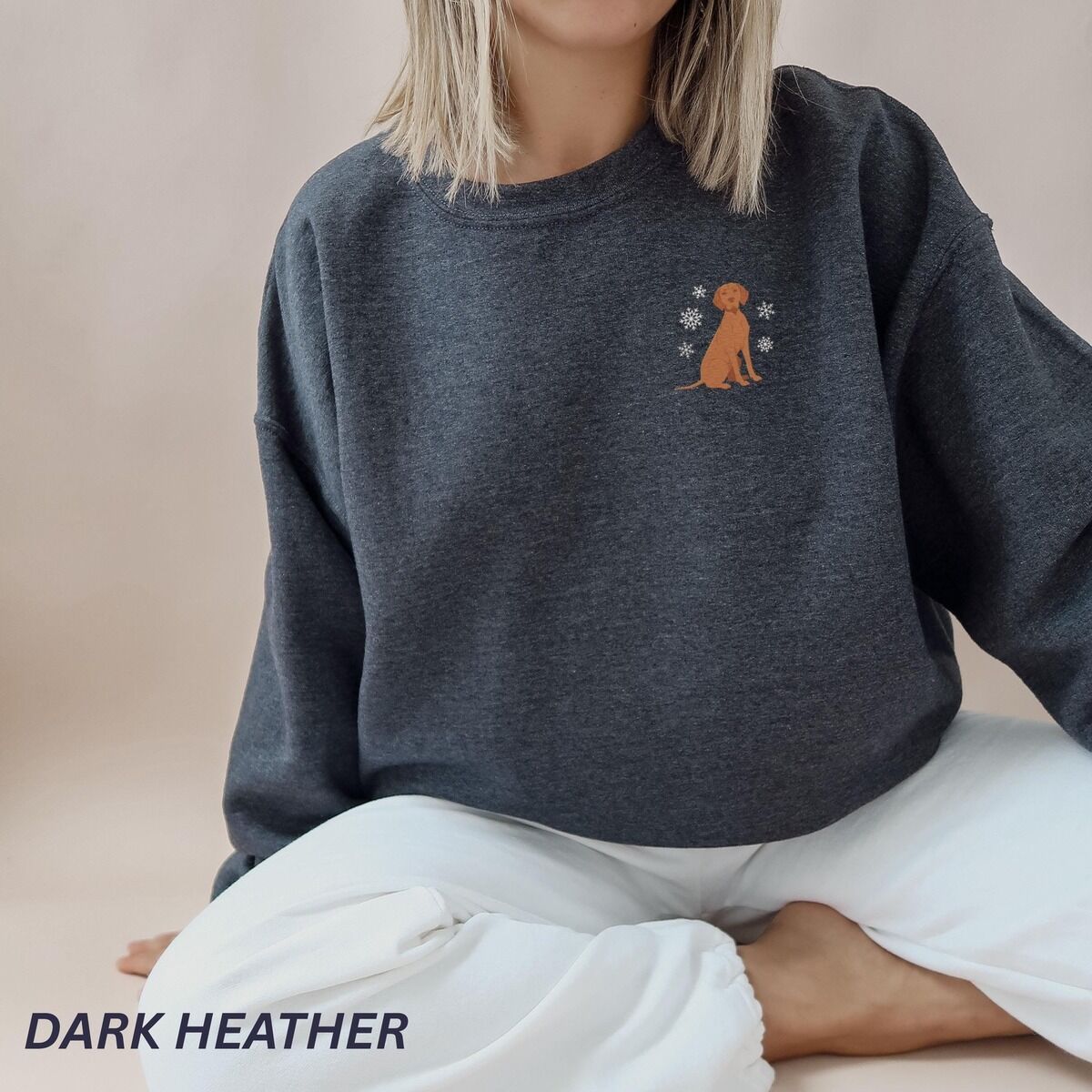 Vizsla Christmas Sweatshirt – Dark Heather Woman sitting in white pants and a Dark Heather crewneck sweatshirt embroidered with a small golden rust vizsla sitting among white snowflakes on the left chest