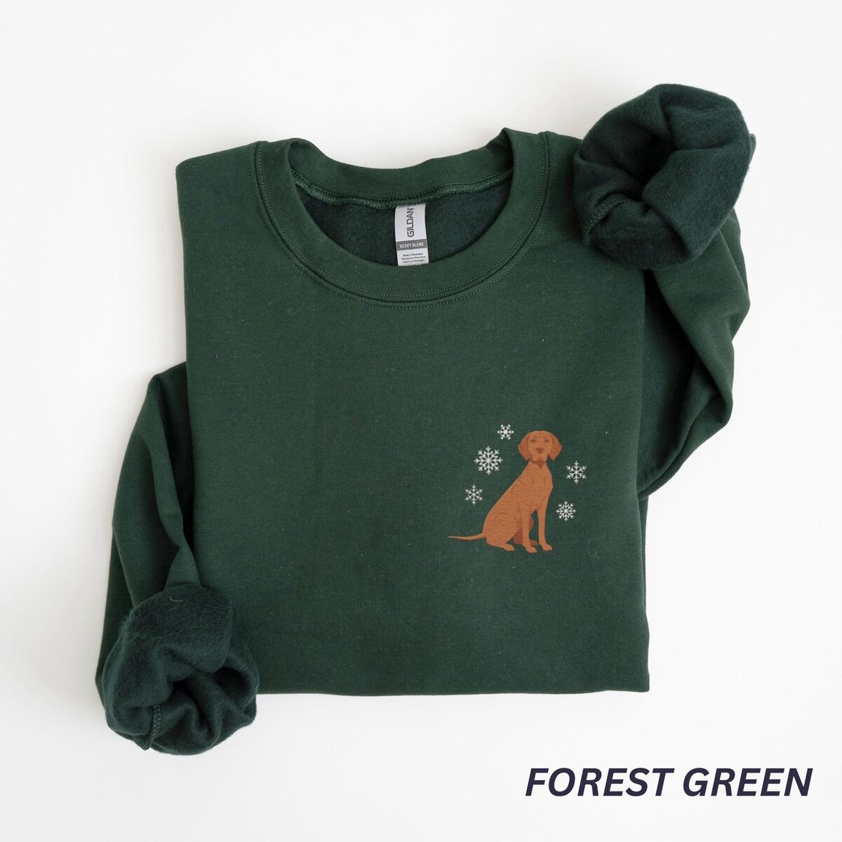 Vizsla Christmas Sweatshirt – Forest Green Forest Green crewneck sweatshirt laid flat on a white background, embroidered with a small golden rust vizsla sitting among white snowflakes on the left chest