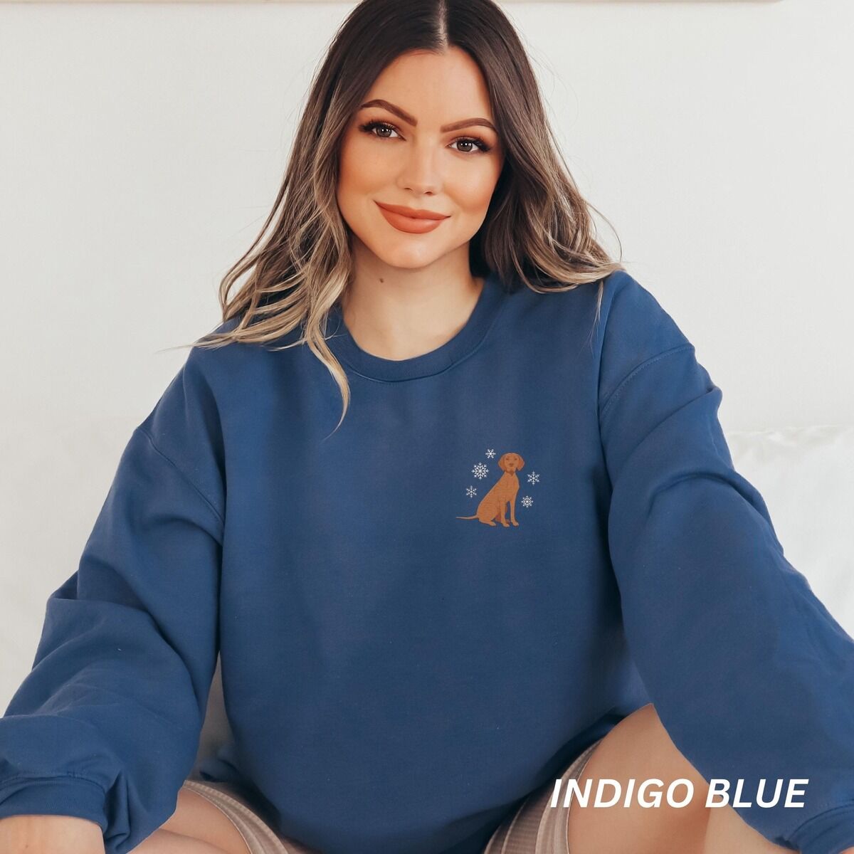 Vizsla Christmas Sweatshirt – Indigo Blue Smiling woman sitting in an Indigo Blue crewneck sweatshirt embroidered with a small golden rust vizsla sitting among white snowflakes on the left chest