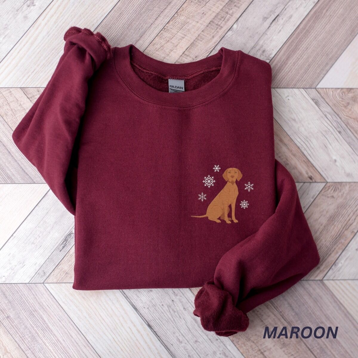 Vizsla Christmas Sweatshirt – Maroon Maroon crewneck sweatshirt laid flat on a herringbone wood floor, embroidered with a small golden rust vizsla sitting among white snowflakes on the left chest