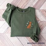 Military Green crewneck sweatshirt laid flat on a herringbone wood floor, embroidered with a small golden rust vizsla sitting among white snowflakes on the left chest
