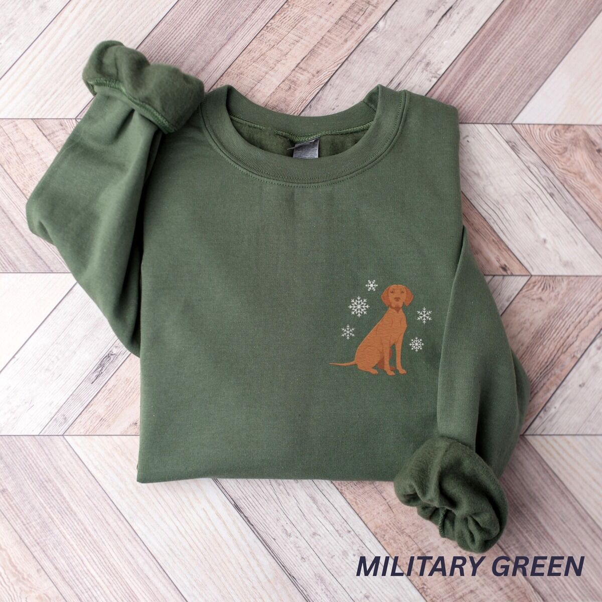 Vizsla Christmas Sweatshirt – Military Green Military Green crewneck sweatshirt laid flat on a herringbone wood floor, embroidered with a small golden rust vizsla sitting among white snowflakes on the left chest
