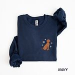 Navy crewneck sweatshirt laid flat on a white background, embroidered with a small golden rust vizsla sitting among white snowflakes on the left chest