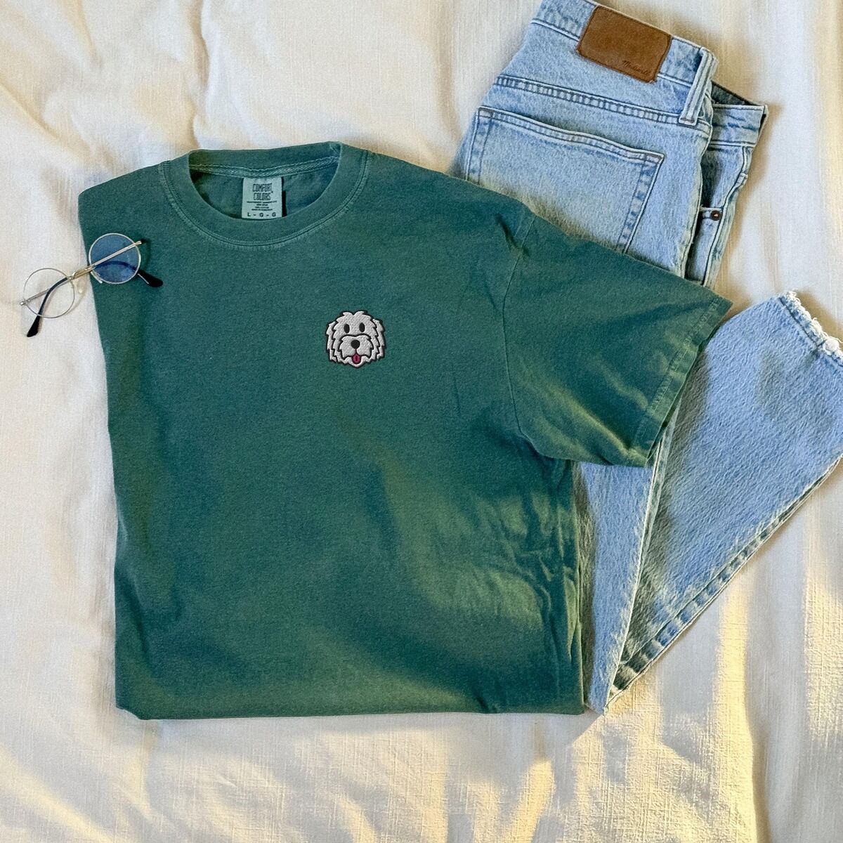 White Goldendoodle Shirt – Blue Spruce Blue Spruce T-shirt laid flat on a white blanket beside jeans and glasses, embroidered with a white cartoon doodle face with a black outline and a pink tongue on the left chest