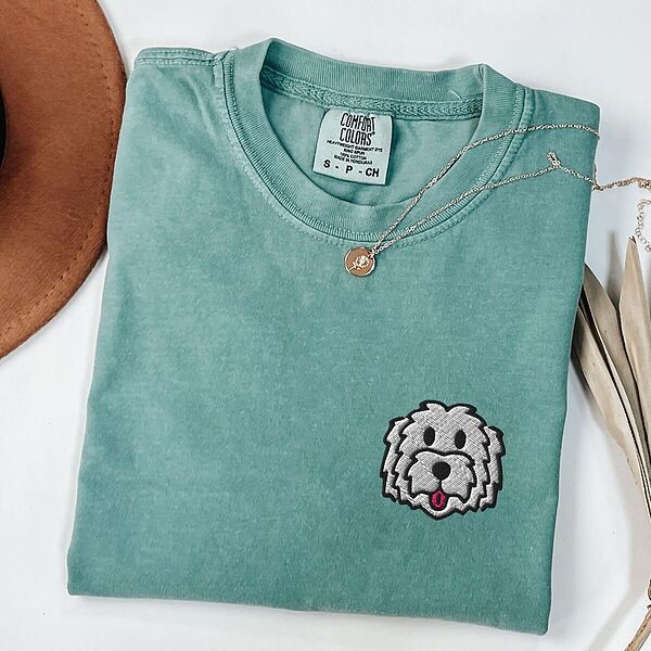 Light Green T-shirt laid flat with a tan hat and a gold necklace, embroidered with a white cartoon doodle face with a black outline and a pink tongue on the left chest