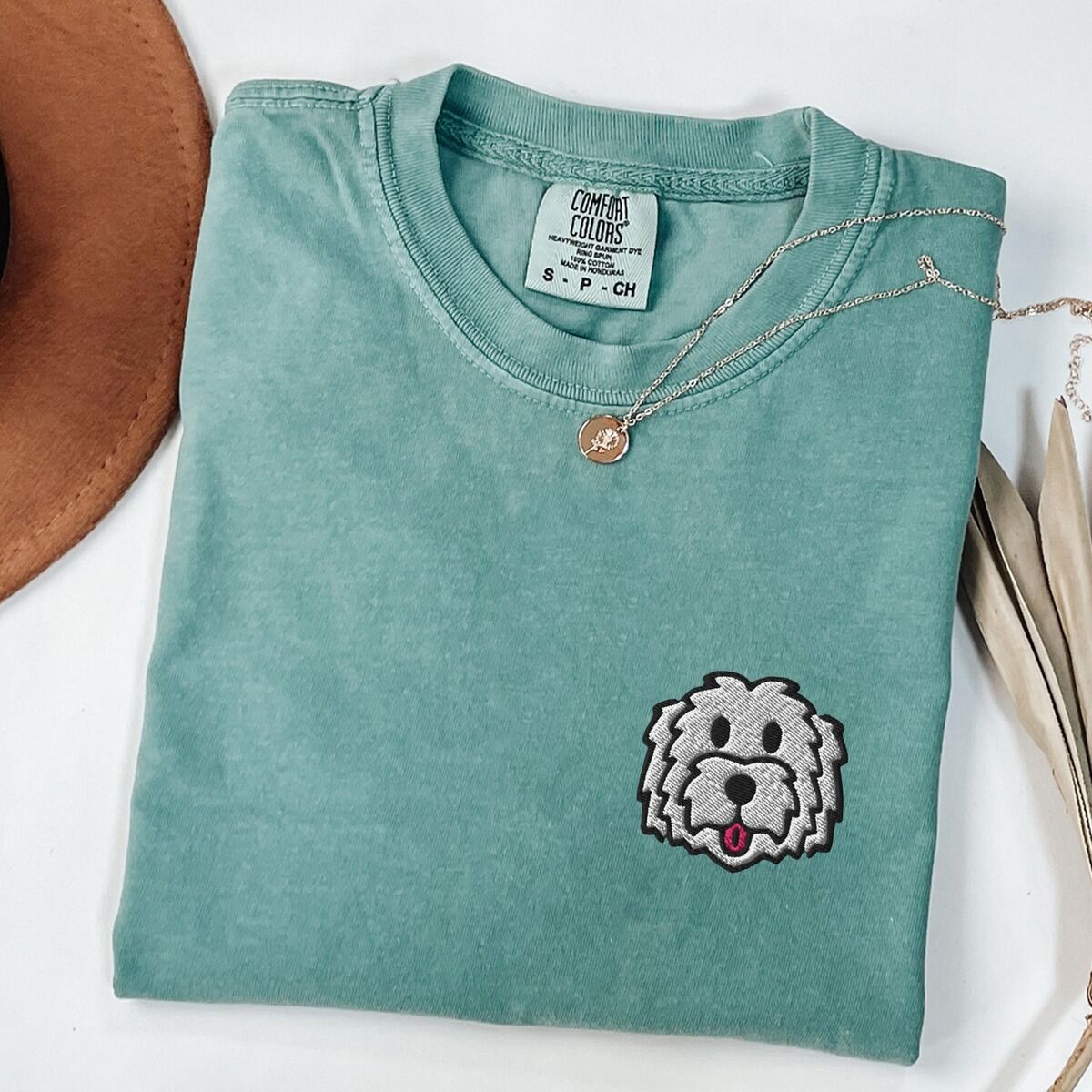 White Goldendoodle Shirt – Light Green Light Green T-shirt laid flat with a tan hat and a gold necklace, embroidered with a white cartoon doodle face with a black outline and a pink tongue on the left chest