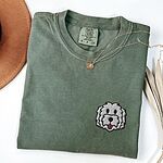 Moss T-shirt laid flat with a tan hat, a gold necklace and dried palm leaves, embroidered with a white cartoon doodle face with a black outline and a pink tongue on the left chest