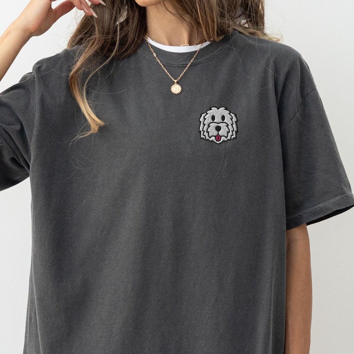 White Goldendoodle Shirt – Pepper Woman with a gold necklace in a Pepper T-shirt embroidered with a white cartoon doodle face with a black outline and a pink tongue on the left chest