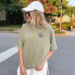 Woman in a white cap on a city street in a light olive T-shirt embroidered with a white cartoon doodle face with a black outline and a pink tongue on the left chest