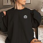 Woman in a Black crewneck sweatshirt embroidered with a white cartoon doodle face with a black outline and a pink tongue on the left chest