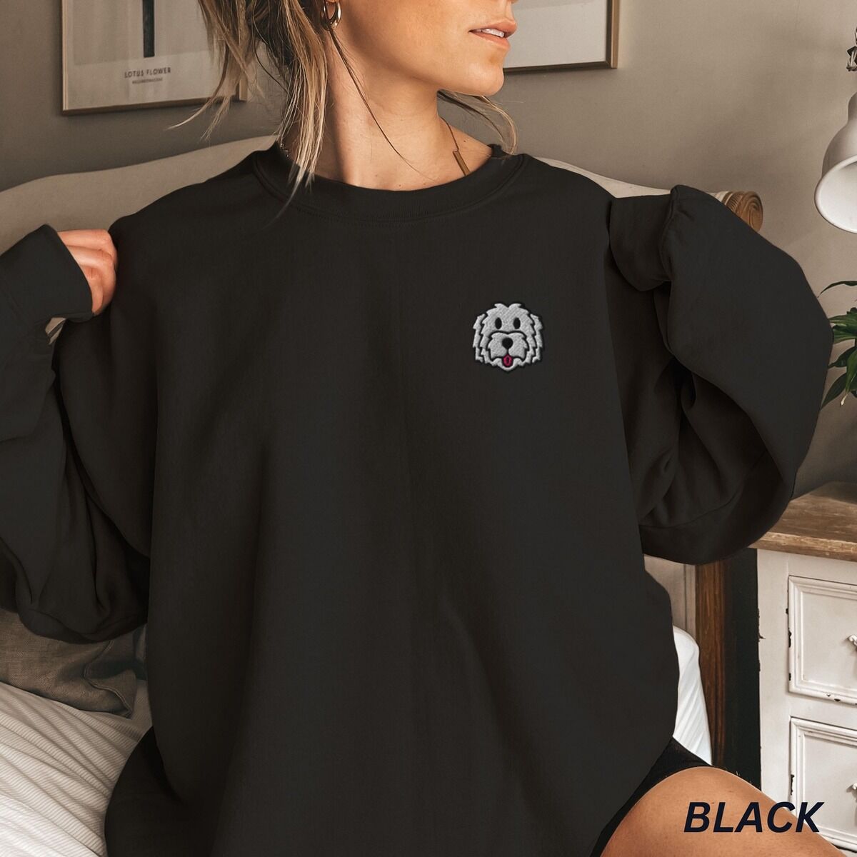 White Goldendoodle Sweatshirt – Black Woman in a Black crewneck sweatshirt embroidered with a white cartoon doodle face with a black outline and a pink tongue on the left chest