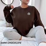 Woman sitting on a bed in a Dark Chocolate crewneck sweatshirt embroidered with a white cartoon doodle face with a black outline and a pink tongue on the left chest