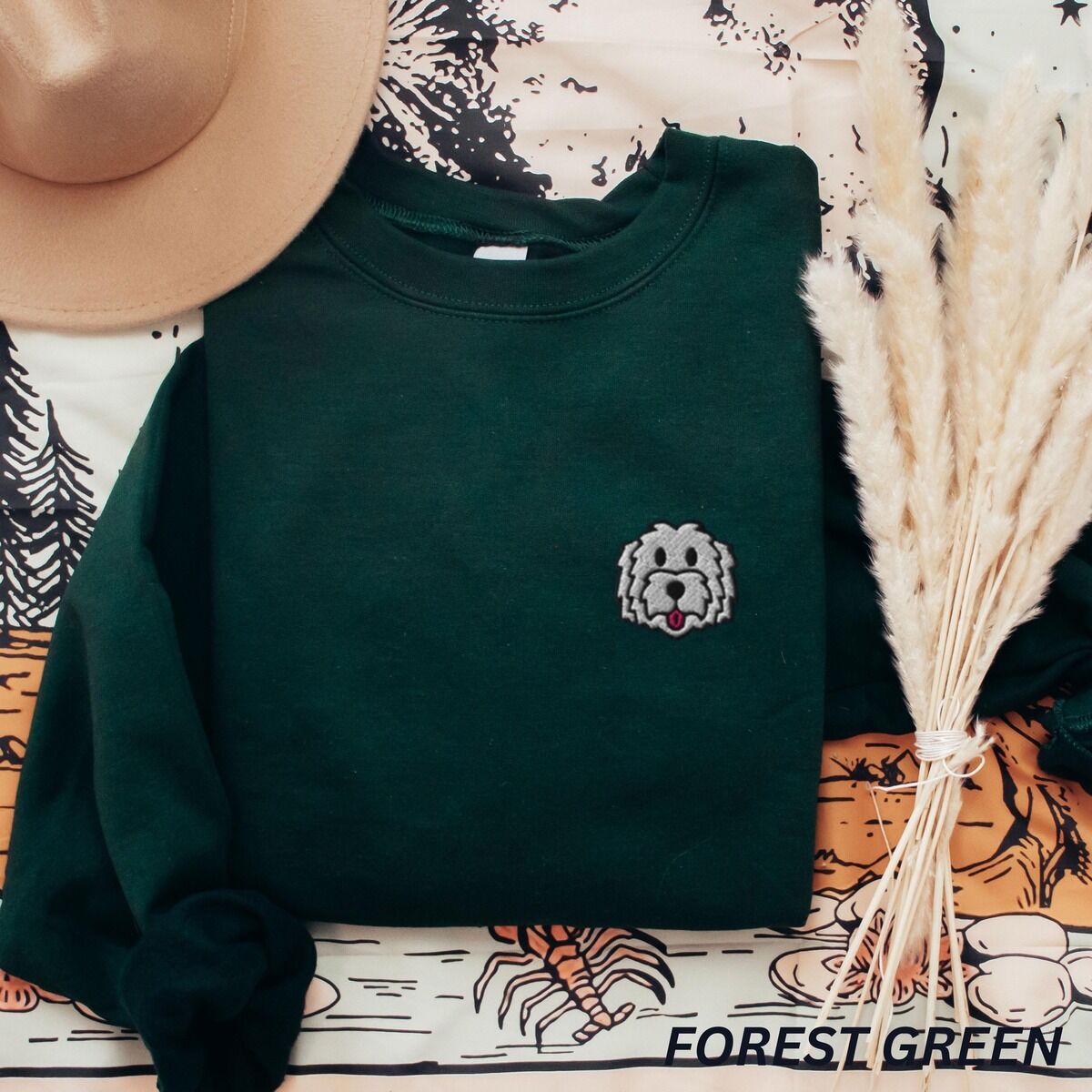 White Goldendoodle Sweatshirt – Forest Green Forest Green crewneck sweatshirt laid flat with a tan hat and dried pampas grass, embroidered with a white cartoon doodle face with a black outline and a pink tongue on the left chest