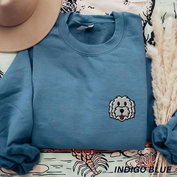 Indigo Blue crewneck sweatshirt laid flat with a tan hat and dried pampas grass, embroidered with a white cartoon doodle face with a black outline and a pink tongue on the left chest