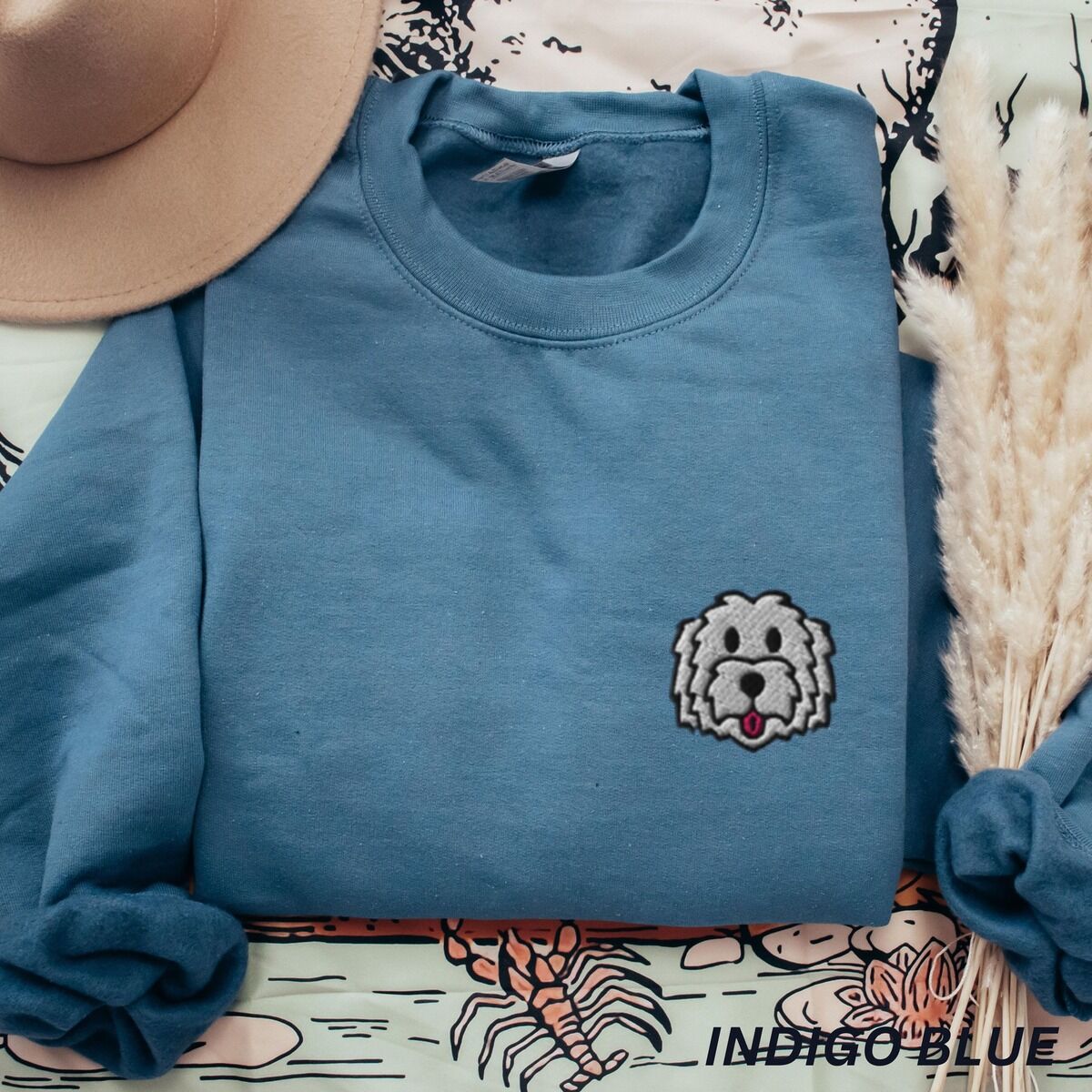 White Goldendoodle Sweatshirt – Indigo Blue Indigo Blue crewneck sweatshirt laid flat with a tan hat and dried pampas grass, embroidered with a white cartoon doodle face with a black outline and a pink tongue on the left chest