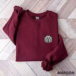 Maroon crewneck sweatshirt laid flat on a herringbone wood floor, embroidered with a white cartoon doodle face with a black outline and a pink tongue on the left chest