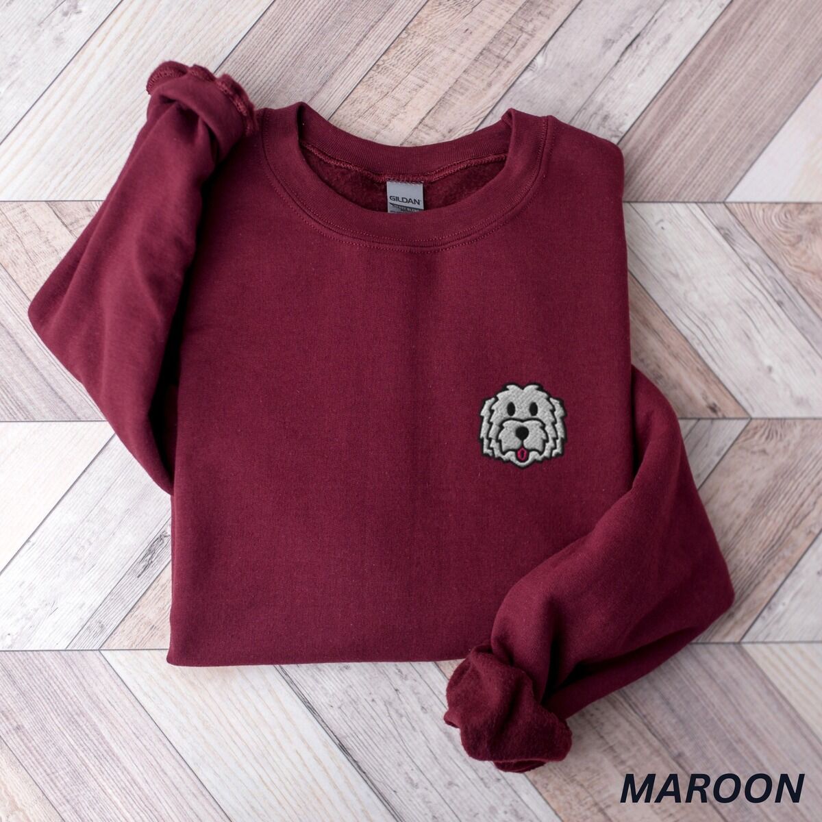 White Goldendoodle Sweatshirt – Maroon Maroon crewneck sweatshirt laid flat on a herringbone wood floor, embroidered with a white cartoon doodle face with a black outline and a pink tongue on the left chest