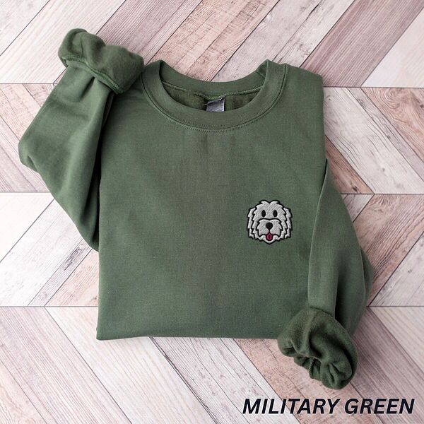 Military Green crewneck sweatshirt laid flat on a herringbone wood floor, embroidered with a white cartoon doodle face with a black outline and a pink tongue on the left chest