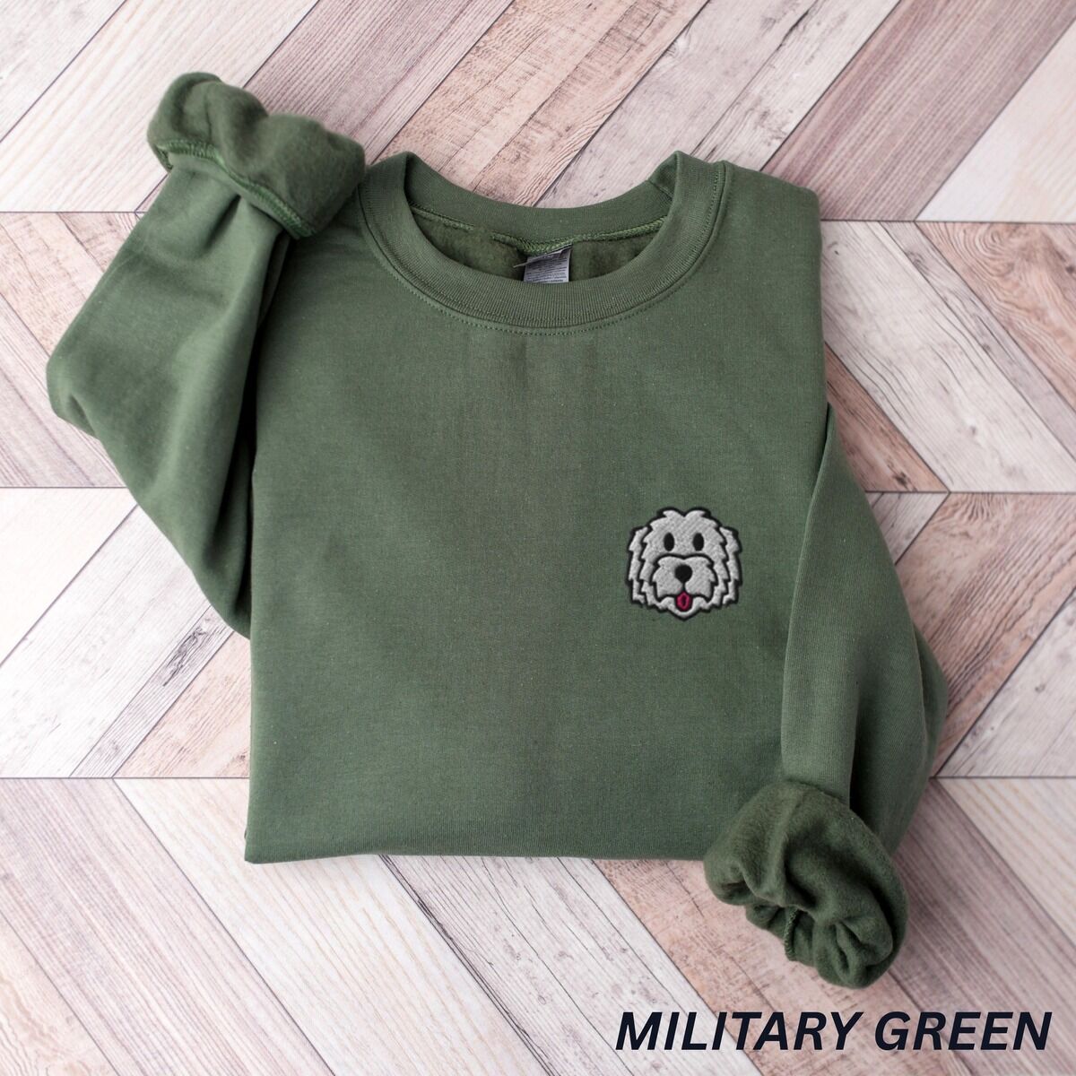 White Goldendoodle Sweatshirt – Military Green Military Green crewneck sweatshirt laid flat on a herringbone wood floor, embroidered with a white cartoon doodle face with a black outline and a pink tongue on the left chest