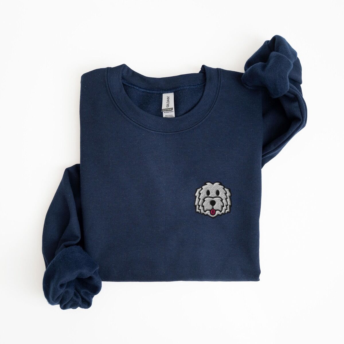 White Goldendoodle Sweatshirt – Navy Navy crewneck sweatshirt laid flat on a white background, embroidered with a white cartoon doodle face with a black outline and a pink tongue on the left chest