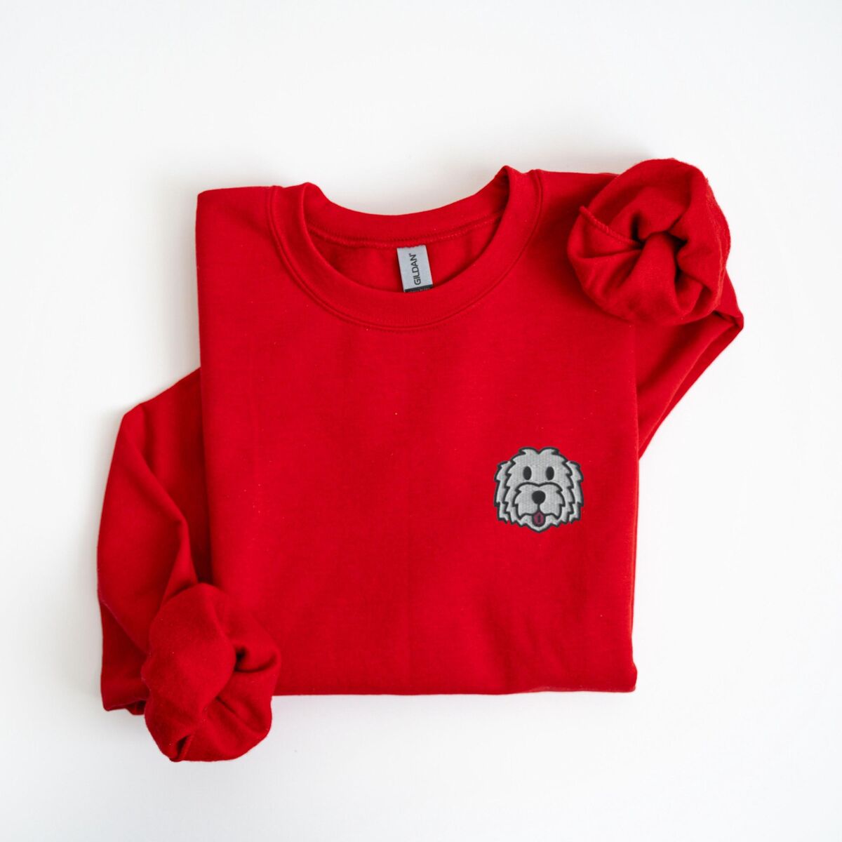 White Goldendoodle Sweatshirt - Red Red crewneck sweatshirt laid flat on a white background, embroidered with a white cartoon doodle face with a black outline and a pink tongue on the left chest