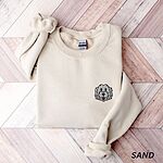 Sand crewneck sweatshirt laid flat on a herringbone wood floor, embroidered with a white cartoon doodle face with a black outline and a pink tongue on the left chest