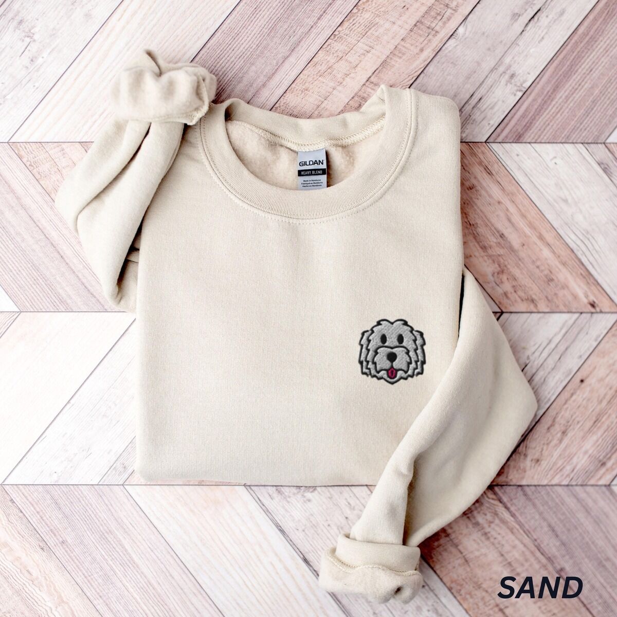 White Goldendoodle Sweatshirt – Sand Sand crewneck sweatshirt laid flat on a herringbone wood floor, embroidered with a white cartoon doodle face with a black outline and a pink tongue on the left chest