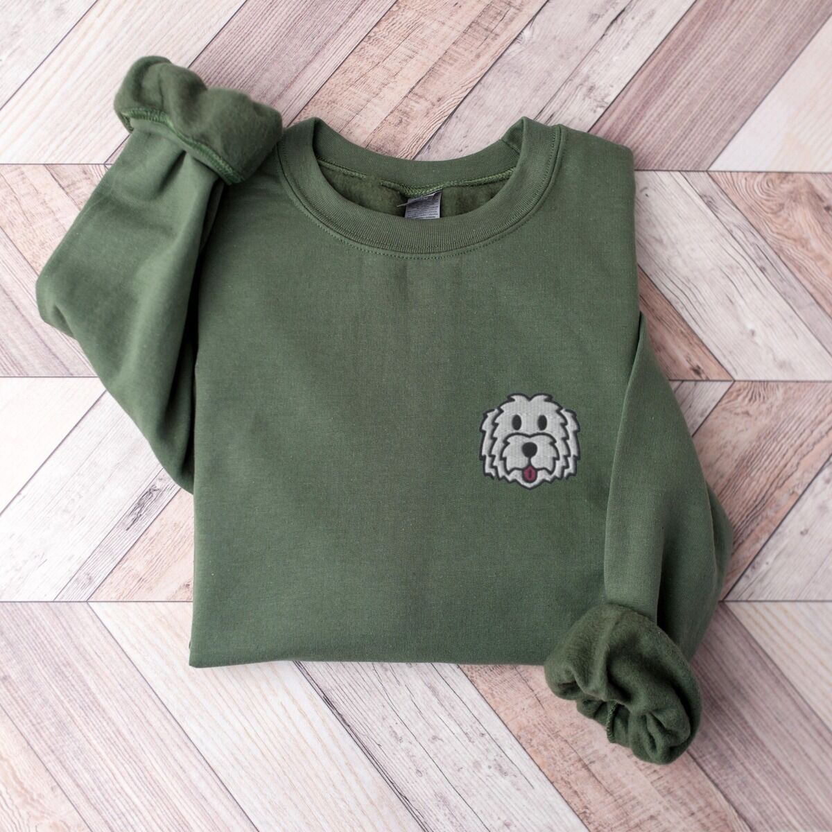 White Goldendoodle Sweatshirt - flat lay Military Green crewneck sweatshirt laid flat on dark wood, embroidered with a white cartoon doodle face with a black outline and a pink tongue on the left chest