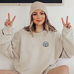 Woman in a beanie making peace signs in a Sand crewneck sweatshirt embroidered with a white cartoon doodle face with a black outline and a pink tongue on the left chest