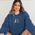 Smiling woman sitting in an Indigo Blue crewneck sweatshirt embroidered with two white labs in Santa hats, one holding a candy cane and one in a long red scarf