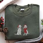 Military Green crewneck sweatshirt laid flat with pine cones and red berries, embroidered with two white labs in Santa hats, one holding a candy cane and one in a long red scarf