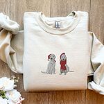Sand crewneck sweatshirt folded on a wood floor beside white roses, embroidered with two white labs in Santa hats, one holding a candy cane and one in a long red scarf
