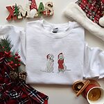 White crewneck sweatshirt laid flat with a plaid scarf, pine and a wooden XMAS sign, embroidered with two white labs in Santa hats, one holding a candy cane and one in a long red scarf