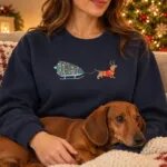 Woman in a Navy wiener dog Christmas sweater sitting by the Christmas tree with her red dachshund