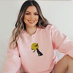 Smiling woman in a Light Pink crewneck sweatshirt embroidered with a black wolf silhouette howling at a yellow full moon with three bats