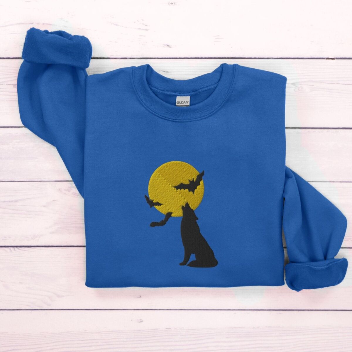 Wolf Howling at the Moon Sweatshirt – Royal Royal crewneck sweatshirt folded on white wooden boards, embroidered with a black wolf silhouette howling at a yellow full moon with three bats