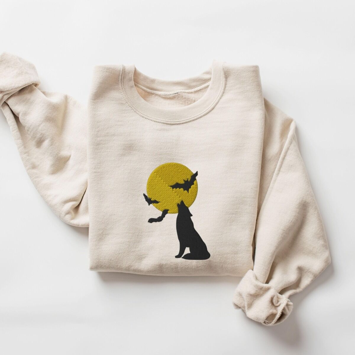 Wolf Howling at the Moon Sweatshirt – Sand Sand crewneck sweatshirt laid flat on a white background, embroidered with a black wolf silhouette howling at a yellow full moon with three bats