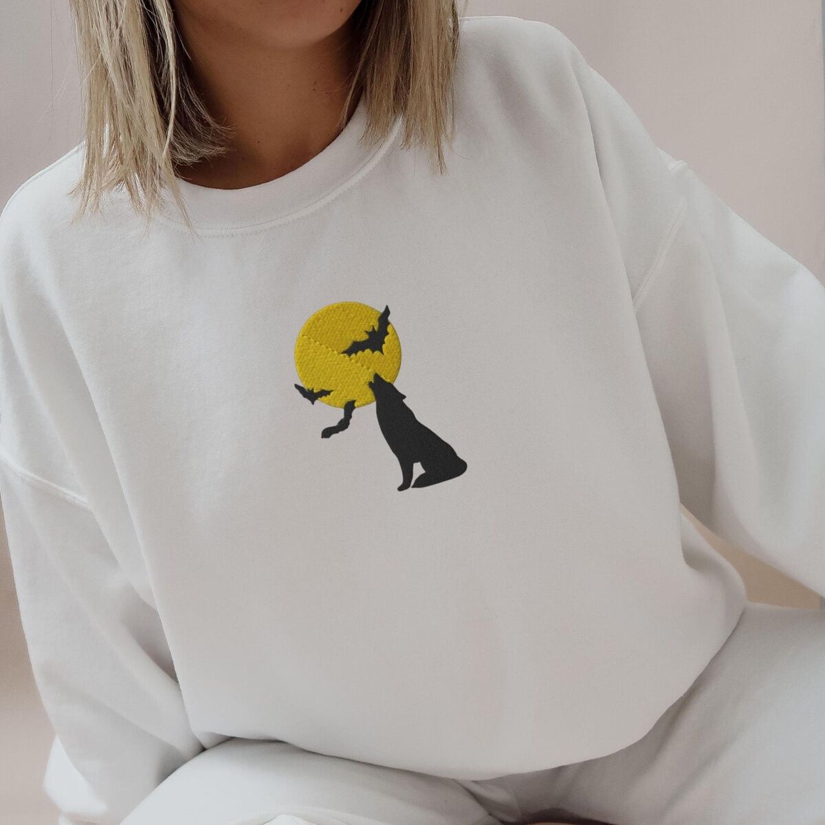 Wolf Howling at the Moon Sweatshirt – White, Worn Woman in a White crewneck sweatshirt embroidered with a black wolf silhouette howling at a yellow full moon with three bats