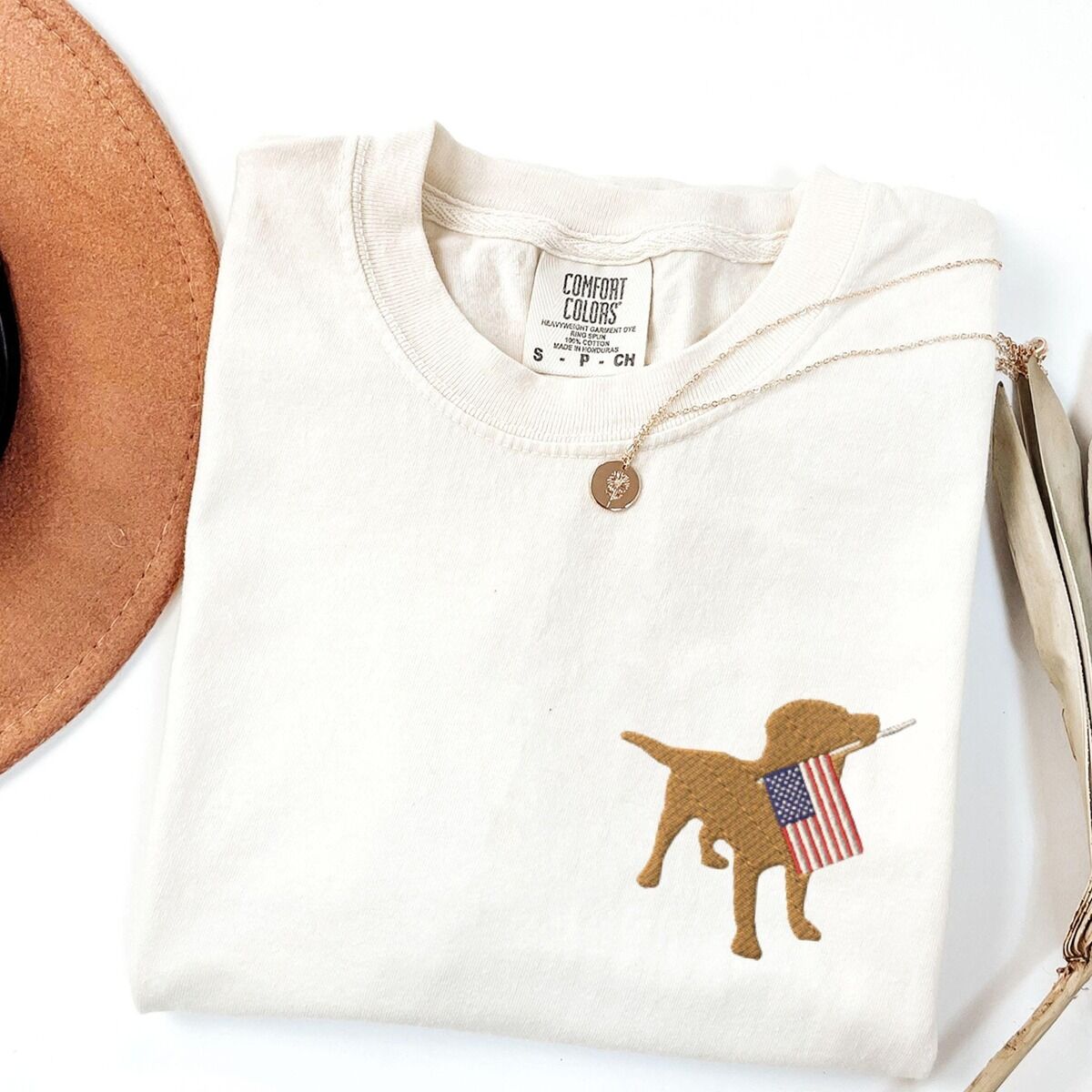 Yellow Lab American Flag Shirt – Ivory Ivory T-shirt folded beside a hat with a small yellow lab holding a stars-and-stripes flag on the left chest