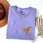 Violet T-shirt laid flat beside a hat and palm leaf, a golden lab silhouette with a USA flag on the left chest