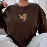 Woman in a Dark Chocolate crewneck sweatshirt embroidered with a yellow lab holding a stars-and-stripes flag across the chest