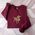 Maroon crewneck sweatshirt laid flat on a wood floor, embroidered in the center with a yellow lab carrying a small flag