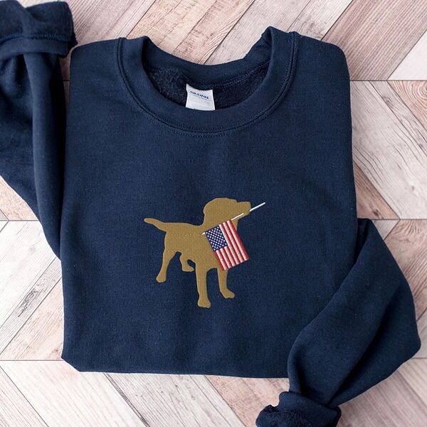 Navy crewneck sweatshirt laid flat on a wood floor, embroidered in the center with a golden yellow lab silhouette carrying a small American flag