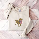 Sand crewneck sweatshirt laid flat on a wood floor, embroidered in the center with a golden yellow lab carrying a stars-and-stripes flag