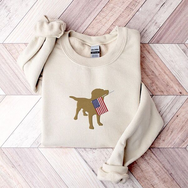 Sand crewneck sweatshirt laid flat on a wood floor, embroidered in the center with a golden yellow lab carrying a stars-and-stripes flag