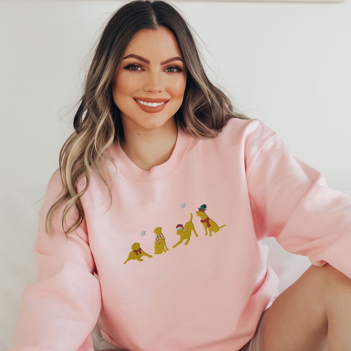Yellow Lab Christmas Sweatshirt - Light Pink Smiling woman sitting in a Light Pink crewneck sweatshirt embroidered with four playful yellow labs in a Santa hat, an elf hat, a red bow and a striped scarf under silver stars